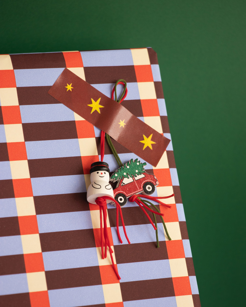 Cadeau verpakt in kleurrijk papier met geblokt patroon, versierd met een gelukspoppetjes van een sneeuwpop en een rode auto met kerstboom.