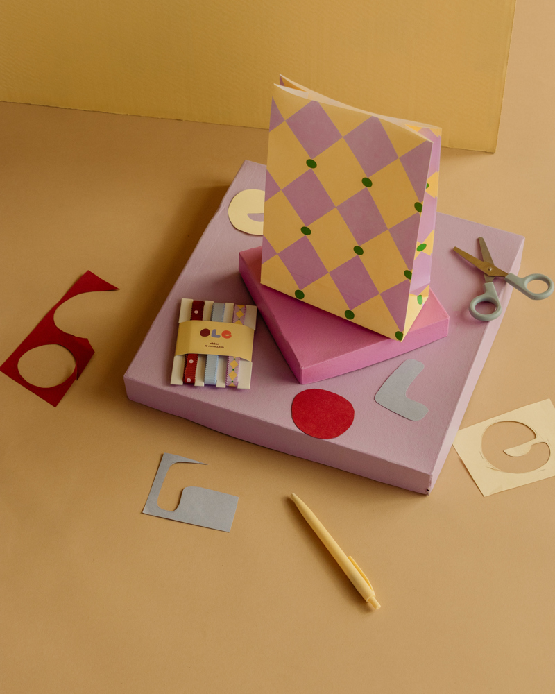 Kleurige papieren materialen, een schaar en een pen liggen op een beige achtergrond met gestapelde paarse en roze dozen.