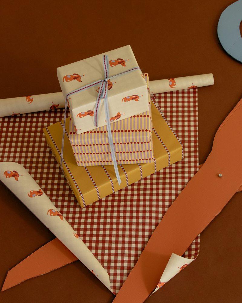Drie ingepakte cadeaus in verschillende kleuren en patronen, liggen op een bruine achtergrond met een stuk inpakpapier ernaast.