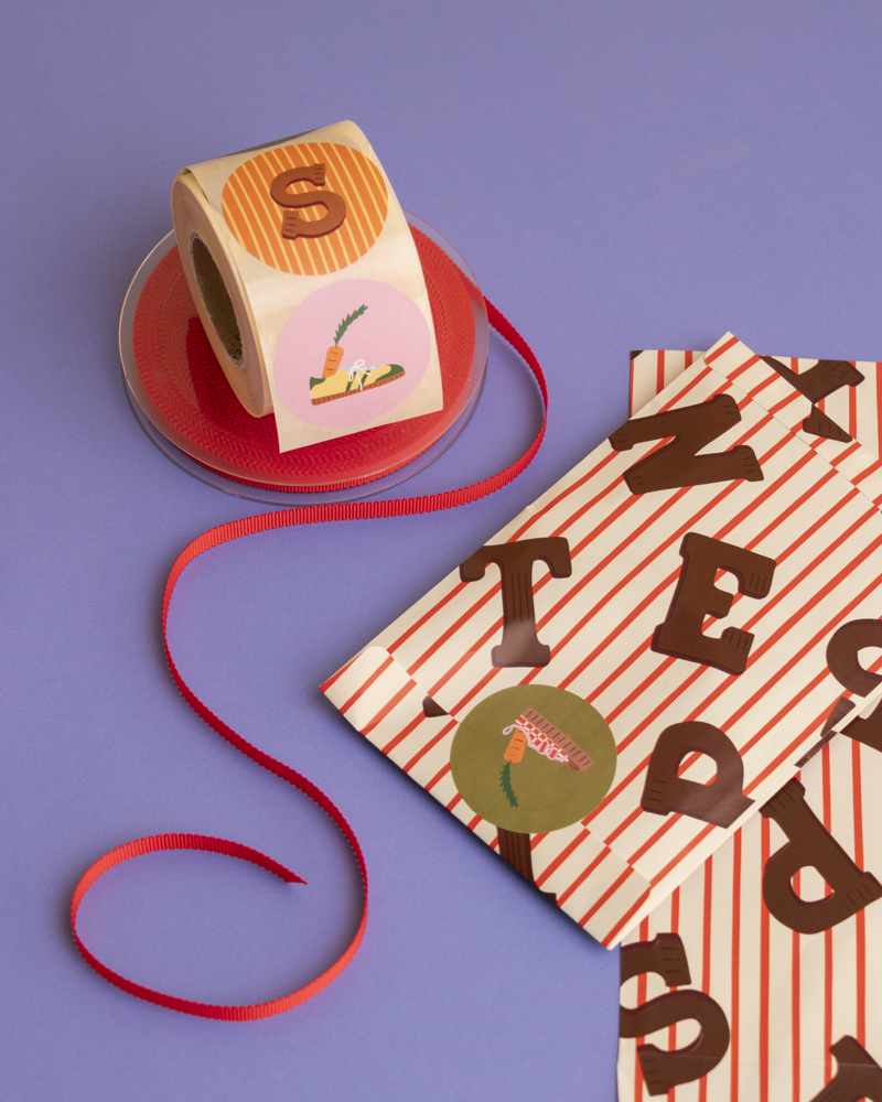 Een rol met stickers, een rood lint en kleurrijke papieren zakjes met letters op een paarse ondergrond.