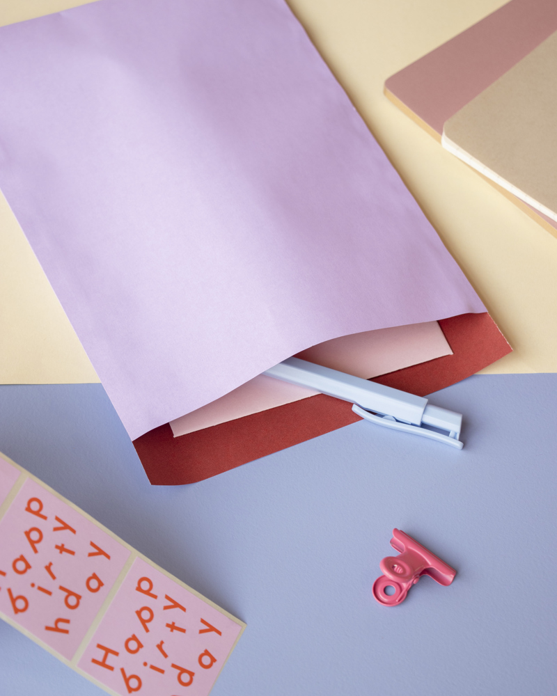 Lila envelop met een blauwe pen, lichtroze en rode vellen papier, een roze clip en een sticker met 'gelukkige verjaardag'.