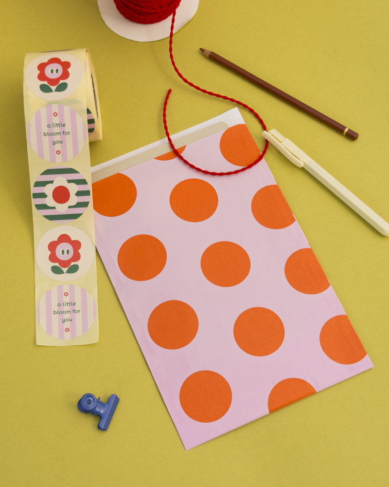 Kleurige achtergrond met een roze envelop met oranje stippen, een rol met bloemstickers, een rood elastiek en een potlood.