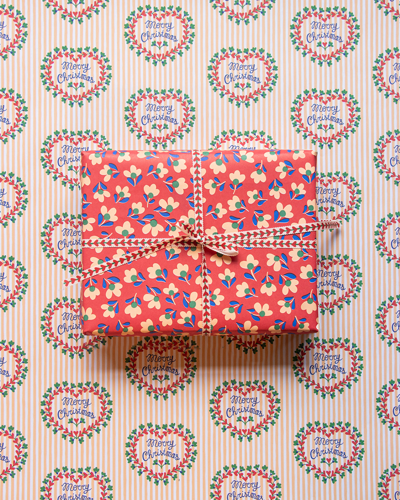 Een cadeau met kleurrijke bloemenprint is ingepakt en staat op een achtergrond met kerstcadeaupapier en "Merry Christmas" in groene kransen.