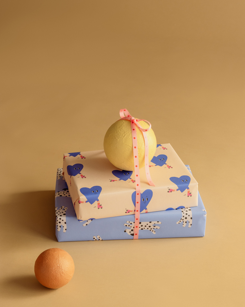 Drie ingepakte cadeaus in verschillende kleuren met een gele bal en een oranje bal op een beige achtergrond.